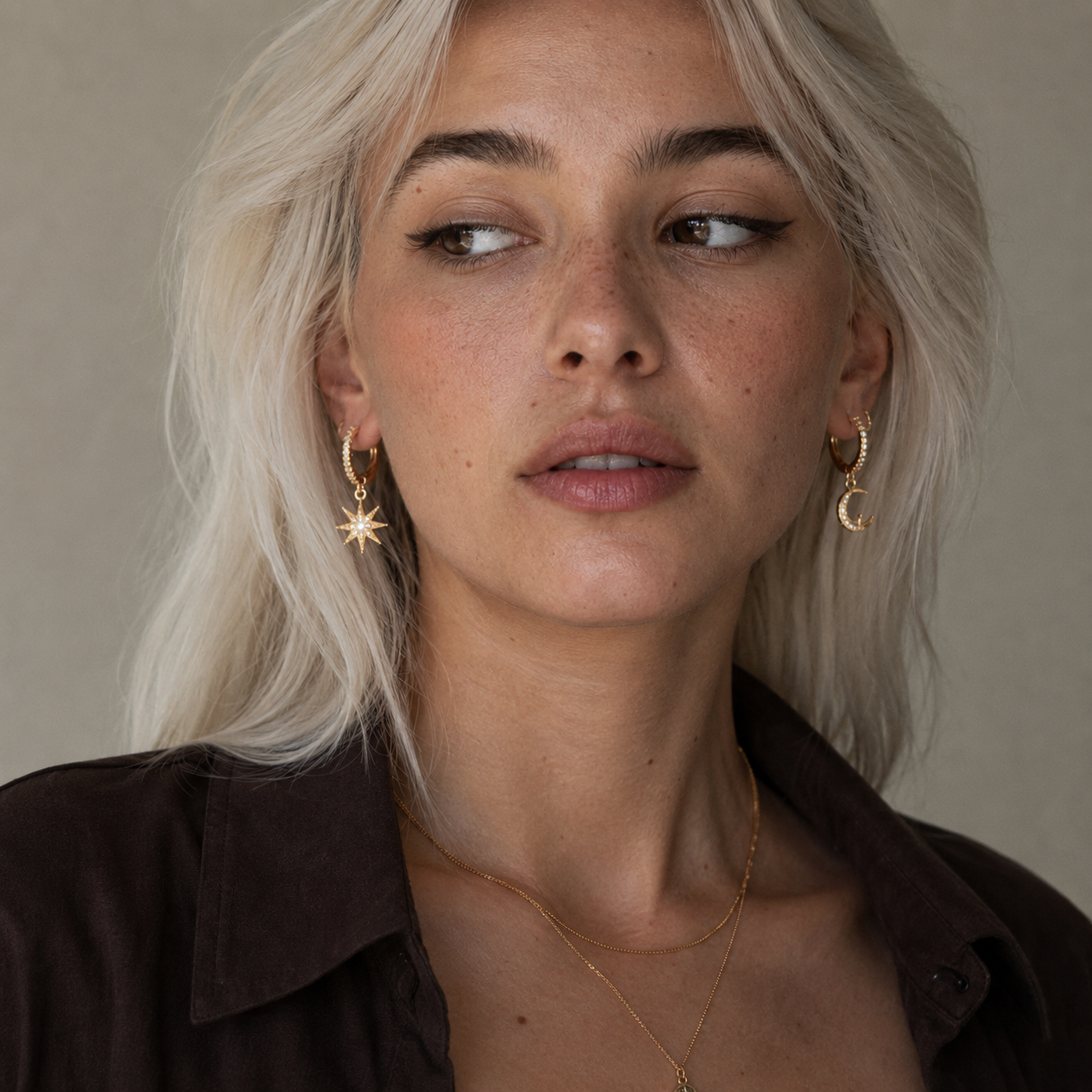 A close-up portrait of a woman with platinum blonde hair wearing gold crescent moon and star hoop earrings, a delicate gold necklace, and a dark brown collared shirt against a neutral beige background.